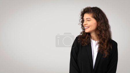 Téléchargez les photos : Femme d'affaires optimiste en costume noir regardant de côté l'espace libre avec un sourire agréable, apparaissant pleine d'espoir et visionnaire, avec un fond gris propre, panorama - en image libre de droit