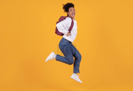 Photo for Joyful young black woman wearing backpack jumping in mid-air against yellow background, happy african american female student expressing excitement and joy, having fun on bright studio backdrop - Royalty Free Image