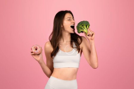 Foto de Enérgica mujer europea feliz en ropa deportiva muerde juguetonamente un floret de brócoli mientras sostiene un donut, que representa opciones de dieta equilibrada contra un alegre telón de fondo rosa, estudio - Imagen libre de derechos