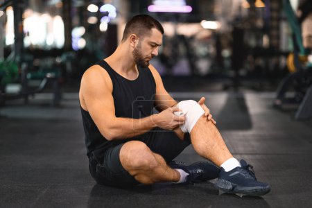 Muscular male athlete attentively wrapping white knee brace around his leg while sitting on gym floor, exemplifying self-care and injury management, got trauma during sport training, copy space