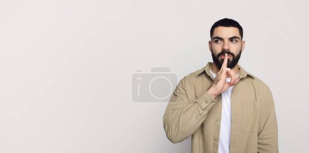 Téléchargez les photos : Sérieux beau jeune homme du Moyen-Orient avec barbe dans occasionnel faire geste shhh avec les lèvres des doigts, regarder l'espace libre, isolé sur fond gris, studio. Nouvelles et potins geste - en image libre de droit