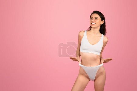 Téléchargez les photos : Une femme européenne joyeuse et vivante aux cheveux foncés rit en détournant les yeux, les mains tirant ludique sur les côtés de son bikini blanc sur un délicieux fond rose, studio - en image libre de droit