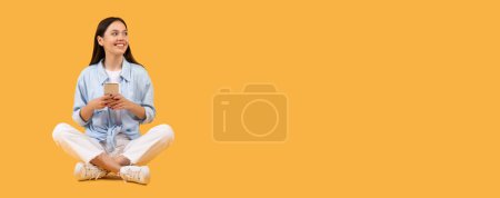 Foto de Jovencita optimista usando smartphone, mirando hacia el espacio libre pensativamente, posando sobre un cálido fondo panorámico naranja, amplio formato, pancarta - Imagen libre de derechos