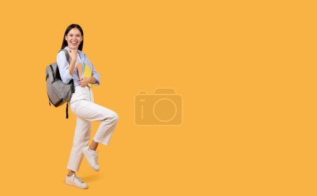 Téléchargez les photos : Étudiante lumineuse et joyeuse avec sac à dos gris tenant des carnets, les poings serrés et souriant, posant sur fond orange, pleine longueur, espace libre - en image libre de droit