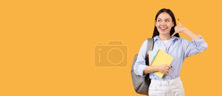 Foto de Estudiante sonriente con mochila, sosteniendo cuadernos y haciéndome llamar gesto de mano, mirando a un lado el espacio libre con expresión reflexiva sobre fondo amarillo brillante - Imagen libre de derechos