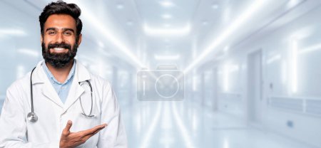Photo for A bearded indian man doctor with a friendly smile is gesturing with an open hand in a hospital setting, suggesting an inviting or explanatory gesture in a clinical environment - Royalty Free Image