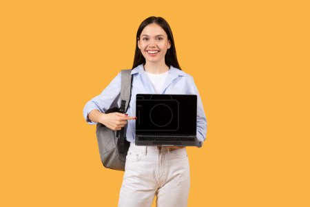 Photo for Confident female student in blue shirt holding an open laptop with blank screen, wearing backpack, presenting educational website against yellow background - Royalty Free Image