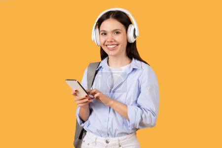 Photo for Smiling female student in casual wear using her smartphone wearing white headphones, carrying grey backpack, standing on yellow background - Royalty Free Image