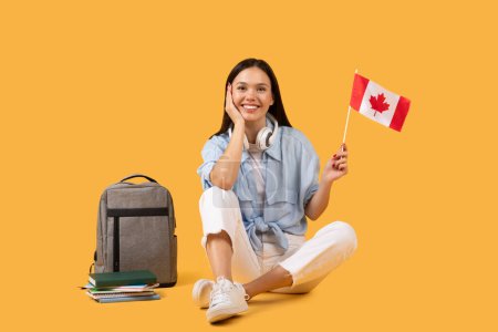Photo for Happy female student holding Canadian flag, wearing headphones, sitting with relaxed pose beside books and backpack on yellow backdrop - Royalty Free Image