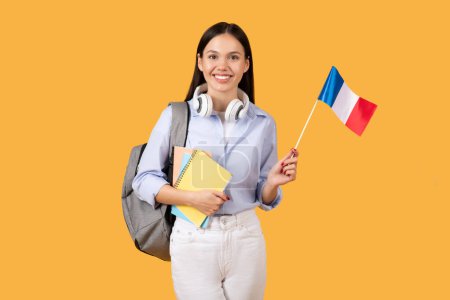 Téléchargez les photos : Étudiante souriante en chemise avec écouteurs blancs, tenant des cahiers et un drapeau français, apprenant des langues étrangères, portant un sac à dos gris sur fond jaune - en image libre de droit