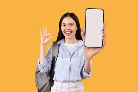 Téléchargez les photos : Jeune femme excitée montrant smartphone avec écran vide tout en faisant signe OK, posant avec sac à dos sur fond jaune, idéal pour l'espace publicitaire éducatif - en image libre de droit