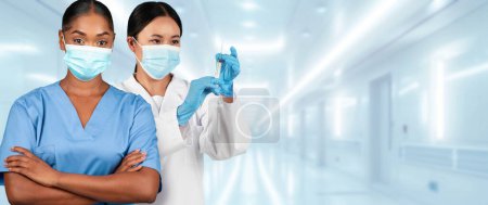 Téléchargez les photos : Deux travailleurs de santé millénaires asiatiques afro-américains dans un cadre clinique, l'un avec les bras croisés dans un gommage bleu et l'autre tenant une seringue, tous deux portant des masques chirurgicaux - en image libre de droit