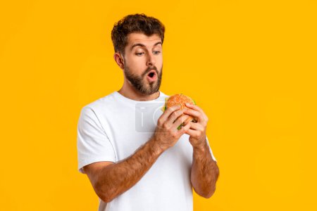 Foto de Retrato de tipo excitado en camiseta blanca sosteniendo y mirando hamburguesa para llevar sobre fondo amarillo, Studio Shot. Hombre hambriento disfrutando sabrosa hamburguesa con queso. Comida chatarra Hábito de comer - Imagen libre de derechos
