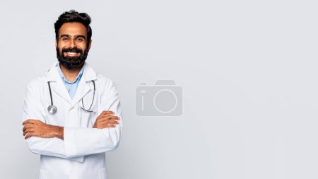 Téléchargez les photos : Médecin indien confiant avec stéthoscope autour du cou et des bras croisés, vêtu d'un blouse de laboratoire, sourit chaudement à la caméra sur fond blanc, espace libre - en image libre de droit