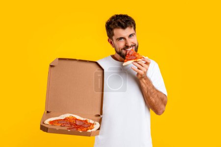 Photo for Happy caucasian man savoring slice of pizza from delivery box, indulges in cheat meal, enjoying junk food in studio setting over yellow background, smiling to camera. Fast food consumption - Royalty Free Image