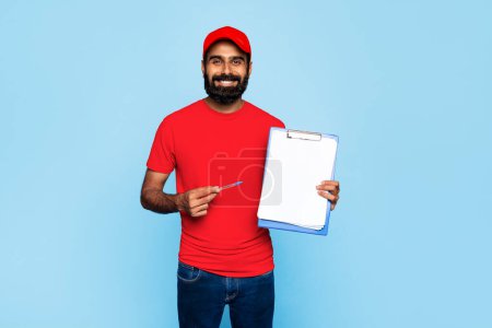 Photo for Cheerful indian delivery man in red uniform holding pen and clipboard with blank paper, ready for signature, standing against vivid blue background - Royalty Free Image
