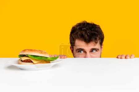 Photo for Caucasian man peeking over table at tempting cheeseburger on plate against yellow studio background. Food cravings for unhealthy eating, nutrition and cheat meal habit concept - Royalty Free Image