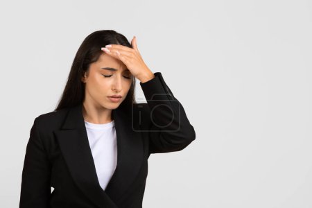 Photo for Professional businesswoman in black suit appears troubled, holding her forehead in gesture of worry, stress, or moment of forgetfulness, grey backdrop - Royalty Free Image