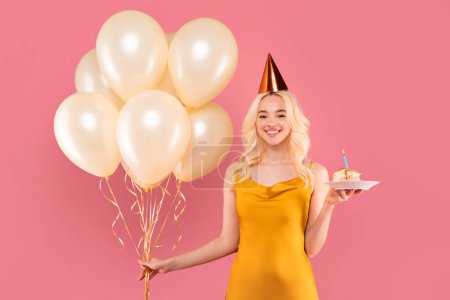 Photo for Smiling young blonde woman in party mood, wearing birthday hat, elegantly holding slice of cake and bunch of cream balloons on pink background - Royalty Free Image