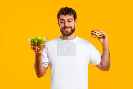 Photo for Food Choice. Smiling Caucasian Young Man Holding Cheeseburger And Healthy Vegetable Salad, Choosing Meal Standing Against Yellow Studio Backdrop. Balance in Nutrition, Junk Food Concept - Royalty Free Image
