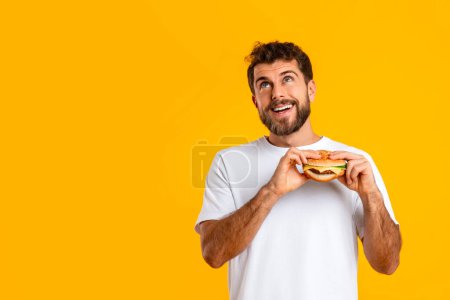 Foto de Tipo hambriento saboreando comida para llevar hamburguesa cerca de espacio de copia en el fondo amarillo, mirando hacia arriba con expresión de ensueño, disfrutando de momentos de placer sobre la dieta, dando en la tentación de la comida chatarra - Imagen libre de derechos