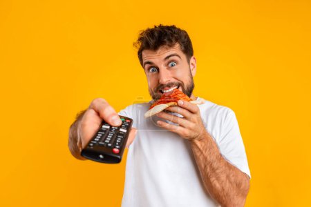 Photo for Young bearded man enjoys slice of pizza while pointing remote control at camera, over yellow studio backdrop. Junk food eater enjoying leisurely moment of indulging in fast food watching television - Royalty Free Image