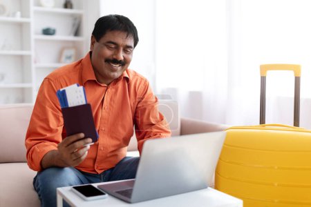 Téléchargez les photos : Réservation de billets en ligne. Homme indien mature excité utilisant un ordinateur portable et achetant tour sur l'agence de voyage Internet, posant près de valise à l'intérieur de la maison moderne, tenant passeport. Concept de plans de vacances - en image libre de droit
