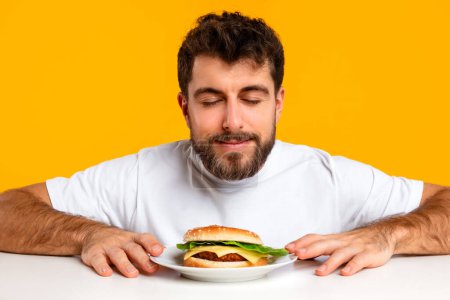 Téléchargez les photos : Jeune homme caucasien ferme les yeux tout en sentant l'arôme d'un hamburger tentant sur l'assiette, assis à table sur fond de studio jaune. Bannière pour contenu nutritionnel et publicité. Réconfort alimentaire - en image libre de droit