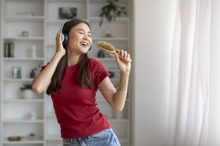 Téléchargez les photos : Femme asiatique joyeuse chantant joyeusement dans la brosse à cheveux comme un microphone, portant des écouteurs, femme coréenne heureuse s'amusant dans le salon lumineux, incarnant le bonheur et le temps libre insouciant - en image libre de droit