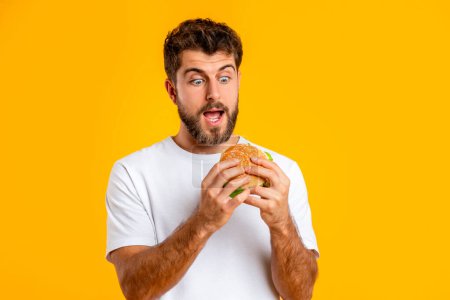 Téléchargez les photos : Portrait de faim excité Millennial Man regardant Burger à bouche ouverte, prêt à mordre la malbouffe savoureuse en studio dans un contexte jaune. Concept d'habitude de manger de la restauration rapide - en image libre de droit