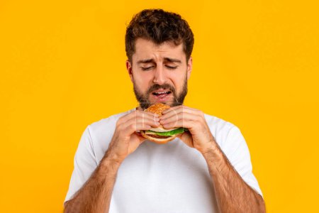 Photo for Unhappy bearded young man eats burger, portraying emotional struggle of balancing healthy diet and temptation of fast food, consuming junk food over yellow backdrop. Studio shot - Royalty Free Image