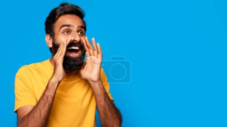 Téléchargez les photos : Enthousiaste homme indien d'âge moyen en chemise jaune criant une annonce avec ses mains taillées autour de sa bouche, regardant vers le côté à l'espace libre sur fond bleu - en image libre de droit