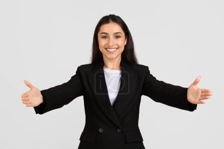 Smiling, professional businesswoman in black suit extends her arms wide in warm, welcoming gesture, inviting cooperation and engagement