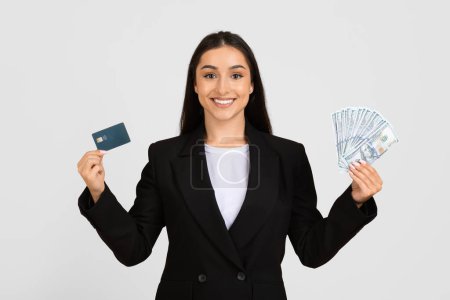 Photo for Happy young businesswoman presenting credit card and fan of US dollar bills, illustrating financial options or purchasing power, isolated on grey background - Royalty Free Image