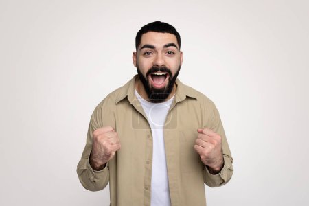 Photo for Happy shocked handsome young latin man winner with beard in casual with open mouth, rise fists up, scream, isolated on gray background, studio. Human emotions, win and success - Royalty Free Image
