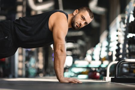 Photo for Focused bearded man doing floor push-ups while training at gym, handsome muscular male athlete showing determination and strength during intense workout session, side view with copy space - Royalty Free Image