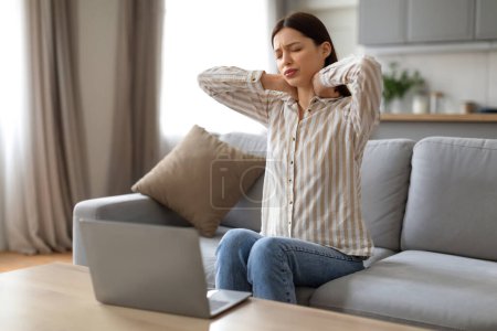 Téléchargez les photos : Jeune femme fatiguée massant son cou, montrant des signes de tension et d'inconfort tout en travaillant à la maison, assise devant son ordinateur portable sur le canapé - en image libre de droit