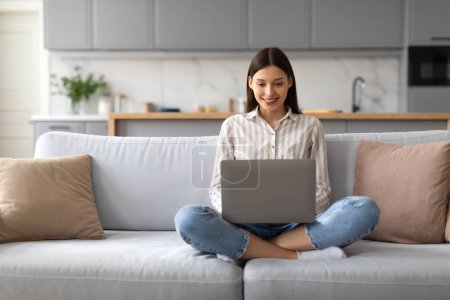Téléchargez les photos : Contenu jeune femme en chemise rayée et jeans travaillant à distance sur ordinateur portable tout en étant assis jambes croisées sur un canapé confortable à la maison, espace libre - en image libre de droit