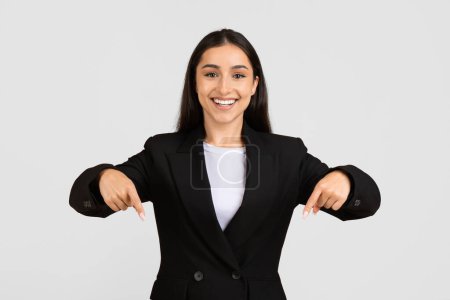 Téléchargez les photos : Femme européenne professionnelle souriante en tenue de travail pointant avec confiance vers le bas avec les deux mains, suggérant un domaine spécifique ou un aspect d'intérêt - en image libre de droit
