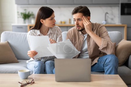 Photo for Young stressed spouses man holding his head in shock and woman showing him document, sitting on couch in front of laptop computer, checking bills - Royalty Free Image