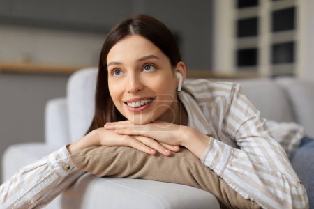 Photo for Young joyful woman with wireless earphones resting on cozy sofa, enjoying music, displaying look of contentment and relaxation in her modern living room - Royalty Free Image