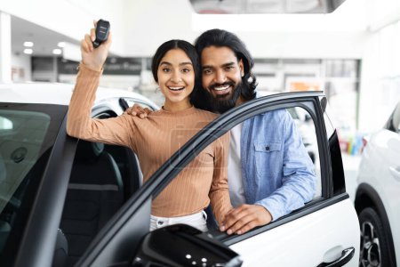 Photo for Happy Owners. Cheerful Indian Spouses Holding Key Of Their New Car And Smiling At Camera While Posing In Dealership Office, Young Eastern Couple Buying Automobile In Modern Showroom - Royalty Free Image