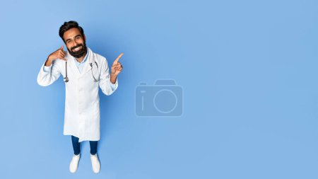 Téléchargez les photos : Joyeux médecin indien d'âge moyen avec stéthoscope me faisant appeler geste de la main et pointant vers l'espace libre, avec un sourire amical sur fond bleu, panorama - en image libre de droit
