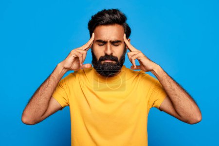 Téléchargez les photos : Homme indien d'âge moyen en chemise jaune avec les yeux fermés, touchant ses tempes, montrant des signes de stress ou de maux de tête graves, sur fond bleu solide - en image libre de droit