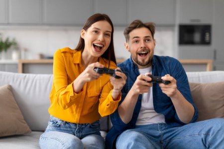 Foto de Joven pareja alegre jugando con entusiasmo a videojuegos, sosteniendo controladores y sentados en el sofá, mostrando competencia lúdica y diversión en el fin de semana en casa - Imagen libre de derechos