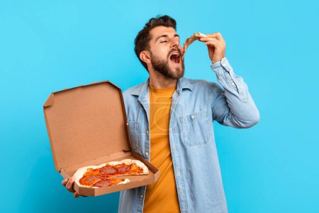 Photo for Caucasian man biting pizza slice with appetite while holding delivery box, standing in studio with blue background, emphasizing concept of comfort eating and unhealthy food choices - Royalty Free Image