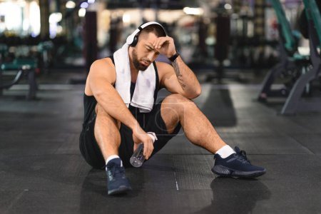 Téléchargez les photos : Athlète masculin fatigué assis sur le sol de la salle de gym avec écouteurs allumés, relaxant après l'entraînement, homme musculaire épuisé en débardeur noir et short essuyant la sueur du front et tenant la bouteille d'eau - en image libre de droit