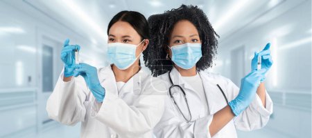 Téléchargez les photos : Deux femmes africaines et européennes diverses, travailleuses de la santé en blouse blanche et gants bleus tiennent des seringues tout en portant des masques chirurgicaux dans un cadre hospitalier clinique, panorama - en image libre de droit
