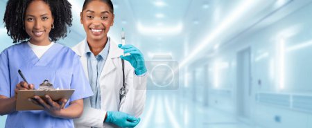 Foto de Dos enfermeras afroamericanas profesionales, una con uniforme azul escrito en un portapapeles y la otra con un abrigo blanco sosteniendo una jeringa, están listas para tareas médicas en un hospital. - Imagen libre de derechos
