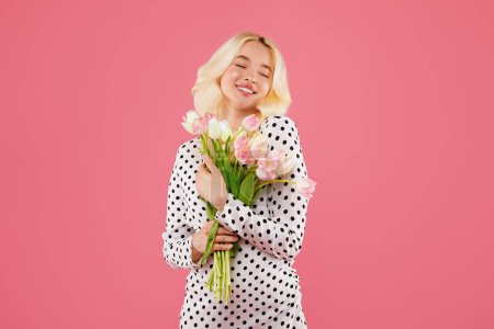 Foto de Joven contenta con los ojos cerrados, abrazando manojo de tulipanes rosados, vistiendo vestido de lunares, celebrando el 8 de marzo sobre fondo rosado - Imagen libre de derechos